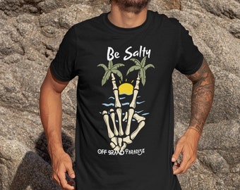 Puede incluir: Una camiseta negra con un gráfico de una mano de esqueleto haciendo un signo de paz con palmeras y un sol detrás. El texto "Be Salty" está por encima del gráfico y "Off Brand Paradise" está por debajo del gráfico.