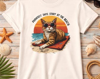 May include: White t-shirt featuring a cartoon cat wearing sunglasses, lounging on a beach towel. The graphic includes a sunset and the text "Purrfect days start at the beach". The shirt is surrounded by beach-themed accessories.