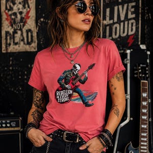 May include: A coral-colored short-sleeve t-shirt featuring a graphic print of a skeleton playing a red electric guitar. The text "REBELLION IN EVERY STROKE" is printed in white below the guitar. The shirt has a crew neckline and rolled sleeves.