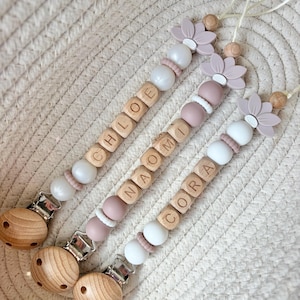 May include: Three dummy clips featuring wooden beads, silicone beads in white and dusty rose, and a flower-shaped silicone accent. Each clip is personalised with the names CHLOE, NAOMI, and CORA, finished with a natural wood clip attachment.