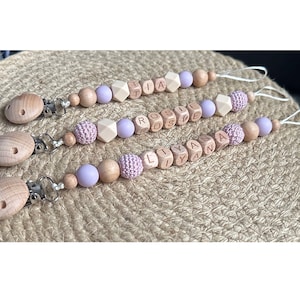 May include: Three wooden baby teethers with clips, each with a different name spelled out in wooden beads. The beads are in shades of light brown and natural wood. The teethers are strung with purple silicone beads.