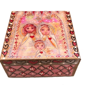 Könnte beinhalten: Eine quadratische Holzkiste mit einer gemalten religiösen Szene auf dem Deckel. Der Deckel zeigt drei Figuren mit roten Herzen vor einem rosa-goldenen Hintergrund. Die Seiten der Box sind mit Herzen und geometrischen Mustern verziert.