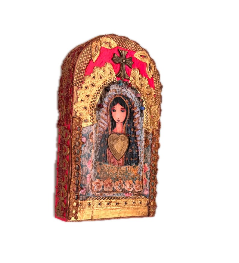 K&ouml;nnte beinhalten: Ein dekorativer, gew&ouml;lbter Wandbehang mit einem zentralen Bild einer Frau mit einem herzf&ouml;rmigen Anh&auml;nger. Das Kunstwerk ist mit Goldfiligran, einem Messingkreuz und einem leuchtend rosa Hintergrund gerahmt. Das St&uuml;ck hat ein strukturiertes, Mixed-Media-Aussehen.