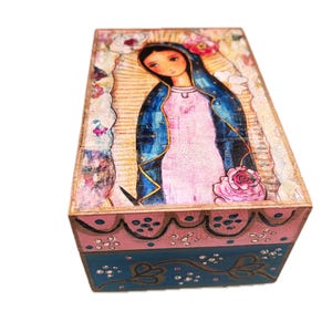 Könnte beinhalten: Eine rechteckige Holzkiste mit einem gemalten Bild einer religiösen Figur. Die Kiste hat ein rosa-blaues Farbschema mit floralen und dekorativen Akzenten. Die Oberseite der Kiste zeigt eine detaillierte Illustration einer Frau mit blauem Schleier und rosa Kleid.