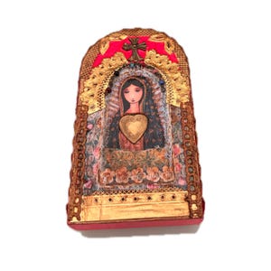 K&ouml;nnte beinhalten: Dekorative religi&ouml;se Ikone mit einem zentralen Bild einer Frau mit einem herzf&ouml;rmigen Emblem. Die Ikone hat eine rote Basis und Oberseite, goldene Akzente und ein Kreuz. Der Hintergrund zeigt florale Muster und goldene Verzierungen.