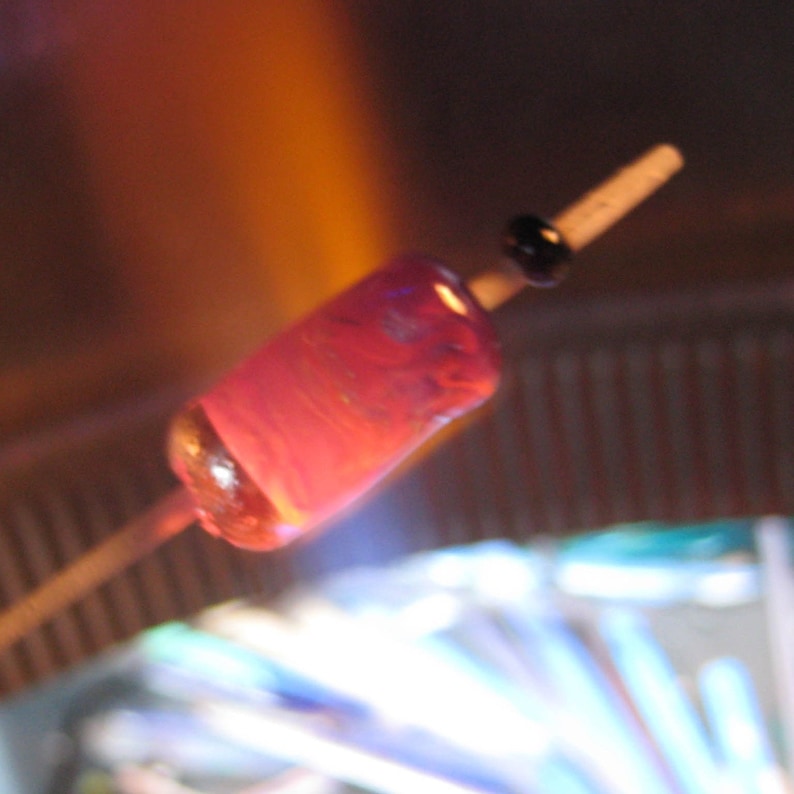 May include: A close-up of a pink glass bead being heated in a flame. The bead is on a metal rod and is being rotated in the flame.