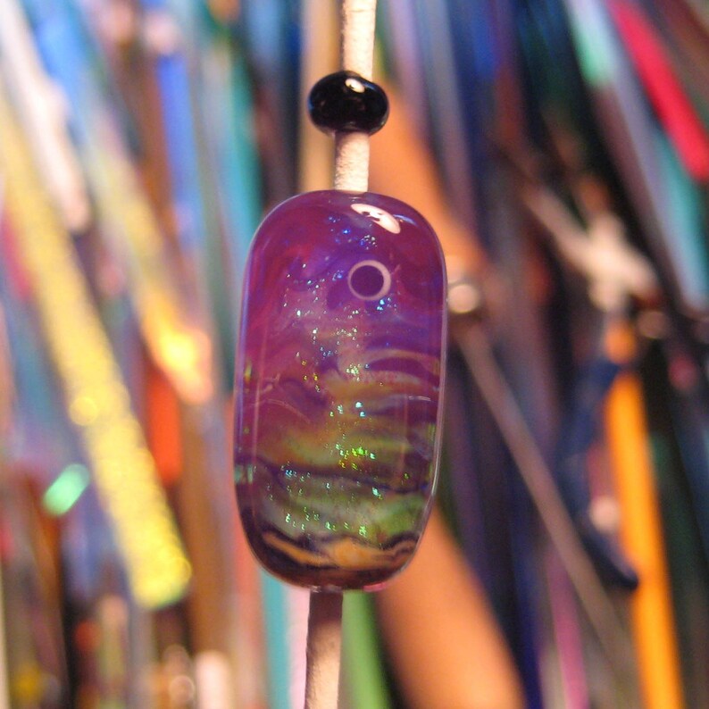 May include: A purple, green, and white glass bead with iridescent glitter. The bead is on a white string and has a small black bead above it.