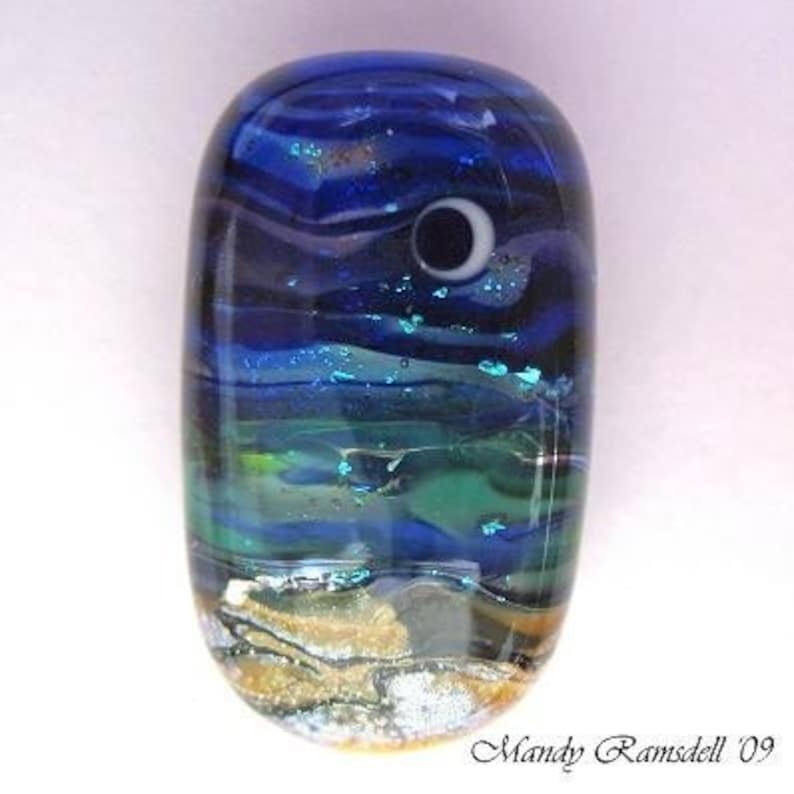 May include: A blue and green glass bead with a white crescent moon design. The bead has a layered effect, with a gold and white layer at the bottom.