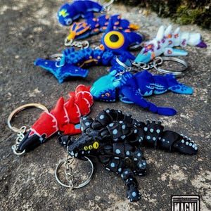 Può includere: Una collezione di portachiavi a forma di creatura articolata in vari colori, tra cui rosso, blu, nero con punti azzurri e bianco con motivi arancioni. Ogni portachiavi presenta un anello in metallo e un corpo flessibile segmentato. Un logo riporta MAGNI MODELS.