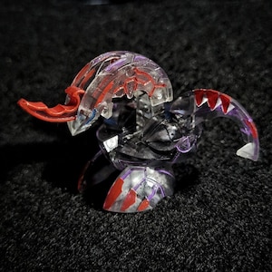 Peut inclure: Une figurine jouet translucide avec des accents rouges et violets. La figurine a un corps segmenté et une tête avec une extension rouge incurvée. Le jouet est principalement noir et transparent, avec des détails rouges et violets.
