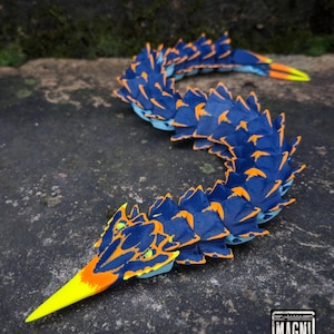 May include: A flexible, articulated toy snake featuring a segmented body with deep blue scales and bright orange accents. The head has a pointed yellow snout and small green eyes. The tail ends in a yellow tip. A logo reads MAGNI MODELS.