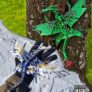 May include: Two articulated dragon figures. One is bright green with black vein-like patterns and red accents, attached to a tree trunk. The other is blue, white, and gold with intricate wing details. A logo in the corner reads MAGNI MODELS.