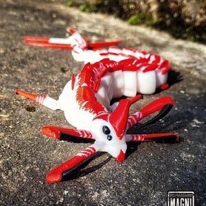 Op de afbeelding: Een 3D-geprint, beweeglijk creatuurmodel in rood en wit, met zwarte accenten op de aanhangsels. Het model ligt op een getextureerd, grijs oppervlak. Het model heeft een gesegmenteerd lichaam en meerdere aanhangsels. Het logo "MAGNI MODELS" is zichtbaar.