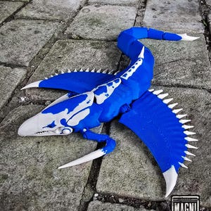 May include: A blue and white model of a plesiosaur, a prehistoric marine reptile, lies on a stone surface. The model features a long neck, flippers, and a detailed design, with white accents and a jagged edge along the flippers.