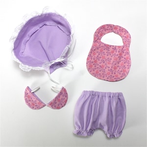 Könnte beinhalten: Ein lilafarbenes Baby-Puppenoutfit mit einer Mütze, einem Lätzchen, einer Latzhose und einem Paar rosa floralen Stoff-Armstulpen.
