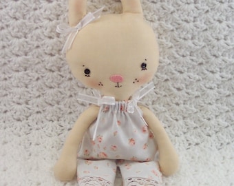 May include: A handmade plush bunny doll with a white dress and pink floral print. The doll has a white bow on its head and black eyes.