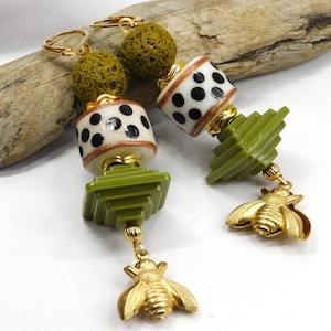 May include: A pair of long, dangling earrings with a green, white, and black color scheme. The earrings feature a green lava rock bead, a white bead with black polka dots, a green geometric bead, and a gold bee charm.