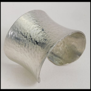 May include: A silver hammered cuff bracelet. The bracelet is wide and has a rounded shape. It is made of a smooth, shiny metal.