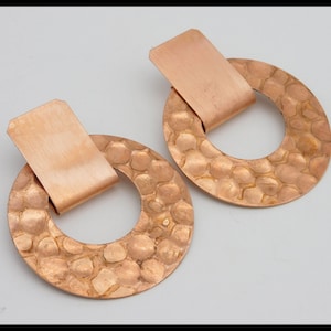 May include: A pair of copper-colored earrings. Each earring features a hammered, textured, circular design with a rectangular accent piece attached at the top. The earrings have a warm, metallic finish, and are displayed against a neutral background.