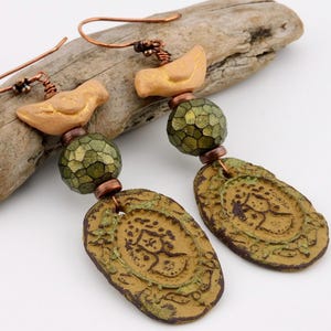 May include: Handmade artisan earrings featuring a bird design. The earrings have copper hooks, tan bird charms, green faceted beads, and oval pendants with an embossed pattern. The color palette includes shades of tan, green, and copper.