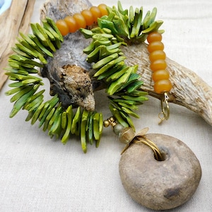 May include: A handcrafted necklace featuring a large, round, gray stone pendant with a central hole. The necklace incorporates green, leaf-shaped beads and amber-colored, rounded beads. The design includes gold-colored metal accents and a clasp.