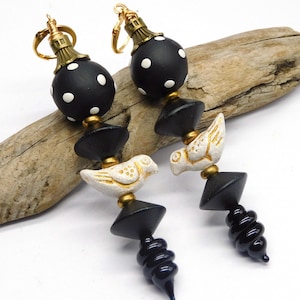 May include: A pair of handcrafted earrings featuring black and white polka dot beads, gold accents, and white bird-shaped charms. The earrings have a unique, artistic design with a dark blue, spiraled bottom.