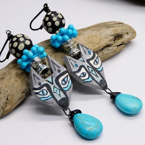 May include: Handmade earrings featuring gray wolf head pendants with blue and white accents. The earrings have turquoise teardrop dangles and black and white polka dot beads. The earrings are connected with black wire and silver-colored metal.