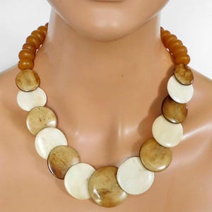 以下が含まれることがあります： クリーム色と茶色の大きな丸いビーズが特徴的なステートメントネックレス。 ネックレスの後ろには、より小さな琥珀色のビーズのストランドがあります。 マネキンに飾られたネックレス。