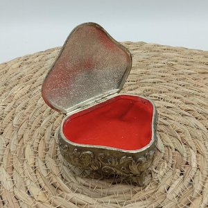 May include: An open, heart-shaped trinket box with a silver-colored exterior and a red velvet interior. The lid is slightly ajar, revealing the plush lining. The box has ornate detailing and rests on small feet.