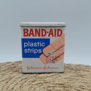 May include: A vintage Band-Aid brand plastic strips tin. The white tin features red and blue text, with an illustration of bandages. The text reads "BAND-AID" and "plastic strips". The bottom of the tin reads "Johnson & Johnson".