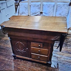 May include: A vintage wooden sewing table with a dark brown finish. The table features a hinged top, three drawers, and decorative carvings on the front panel. The table is supported by four legs and has a drop-leaf extension.