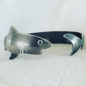 May include: A silver shark cuff bracelet with a black gemstone eye. The bracelet is open and has a textured finish.