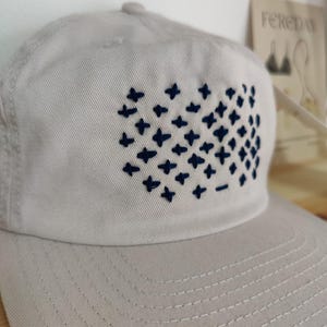 Puede incluir: Una gorra de b&eacute;isbol gris claro con un dise&ntilde;o bordado azul marino de una cuadr&iacute;cula de signos m&aacute;s.