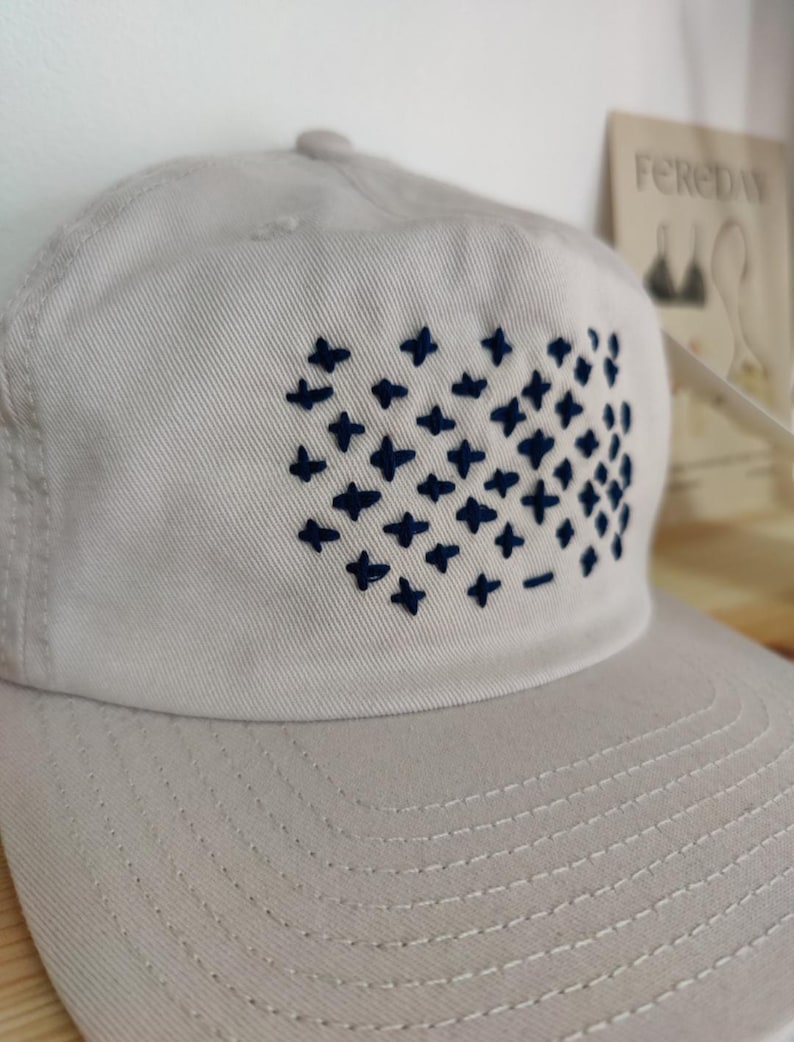 Puede incluir: Una gorra de b&eacute;isbol gris claro con un dise&ntilde;o bordado azul marino de una cuadr&iacute;cula de signos m&aacute;s.