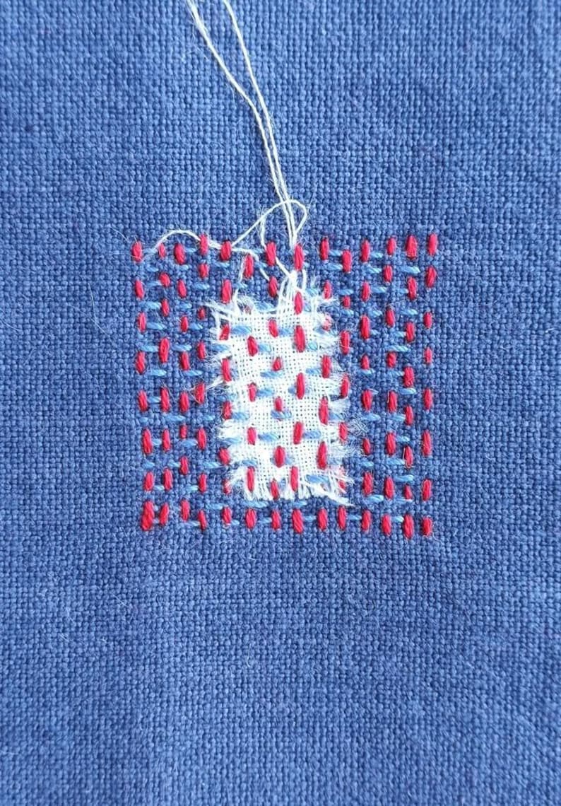 Puede incluir: Primer plano de una tela azul con un cuadrado bordado en rojo y blanco. El bordado es un patr&oacute;n simple de puntadas rojas y blancas.