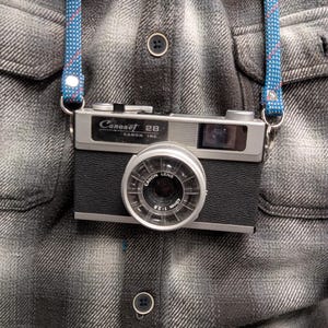 Könnte beinhalten: Eine Vintage Canonet 28 Kamera mit einem schwarzen und silbernen Gehäuse und einem blau-rot gemusterten Gurt. Das Kameraobjektiv ist mit "CANON LENS 40mm 1:2.8" beschriftet. Die Kamera hängt an einem grau karierten Hemd.