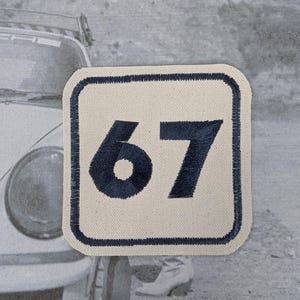 May include: A square patch featuring the number 67 in navy blue embroidery on a cream fabric. The patch has a navy blue border. The background is a blurred black and white image of a vintage car.