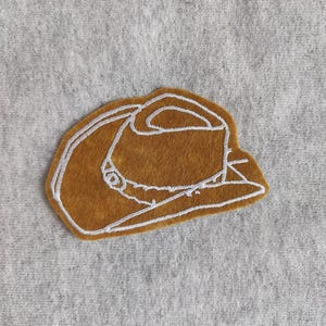 May include: A brown felt cowboy hat with white stitching, embroidered on a gray fabric background.