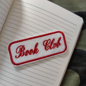 May include: A white rectangular patch with red trim and the words "Book Club" embroidered in red script. The patch is placed on an open notebook with lined pages. The background is a camouflage pattern.