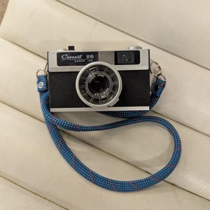 May include: A vintage Canonet 28 camera. It has a black body, silver top, and a blue and red patterned strap. The camera lens is visible, and the text "Canonet 28" is printed on the top. A classic photography item.