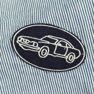 May include: Close-up of a blue and white striped fabric with a navy blue oval patch. The patch features a white outline of a classic car. The car is a coupe with visible wheels and windows. The fabric has vertical stripes.