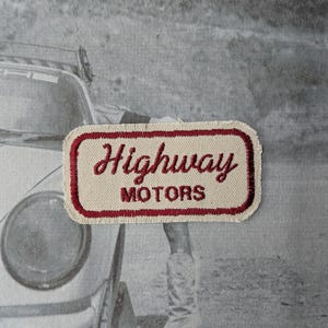 May include: A rectangular patch with the words "Highway Motors" embroidered in red on a cream background, framed by a red border. The patch is set against a black and white image of a car and a person wearing cowboy boots.