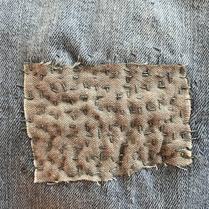 May include: A close-up of a denim fabric with a brown patch sewn on. The patch is made of a rougher fabric and has been stitched with a running stitch in a grid pattern.