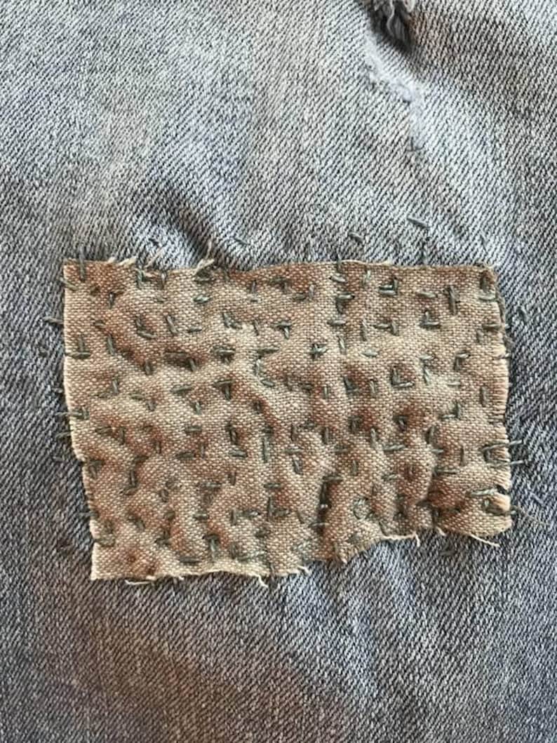 May include: A close-up of a denim fabric with a brown patch sewn on. The patch is made of a rougher fabric and has been stitched with a running stitch in a grid pattern.