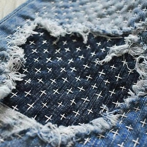 Puede incluir: Primer plano de un par de pantalones vaqueros azules con una zona desgastada que se ha reparado con puntadas blancas en punto de cruz.