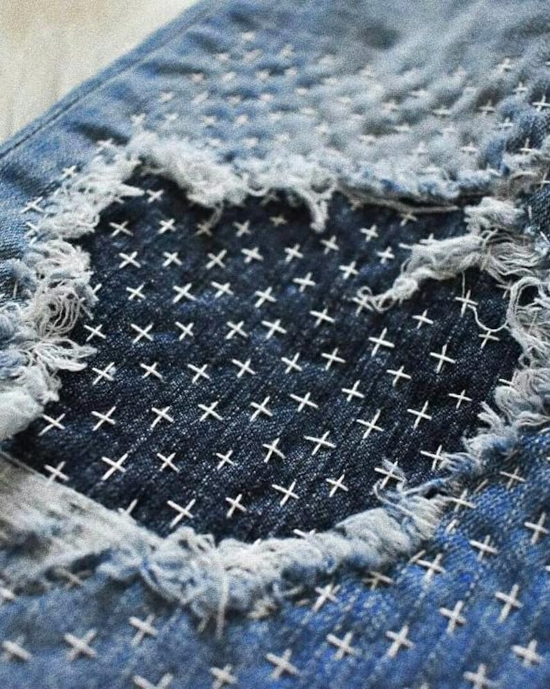 Puede incluir: Primer plano de un par de pantalones vaqueros azules con una zona desgastada que se ha reparado con puntadas blancas en punto de cruz.