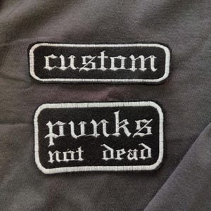 以下が含まれることがあります： 白いステッチが施された黒い布のパッチが2つ。上のパッチにはゴシック体で「custom」と書かれている。下のパッチにはゴシック体で「punks not dead」と書かれている。