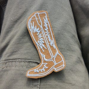 May include: A brown embroidered patch of a cowboy boot with white stitching.