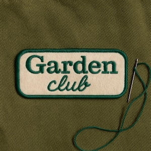 May include: A white felt patch with green embroidered text that reads "Garden club". The patch is on a green fabric background.