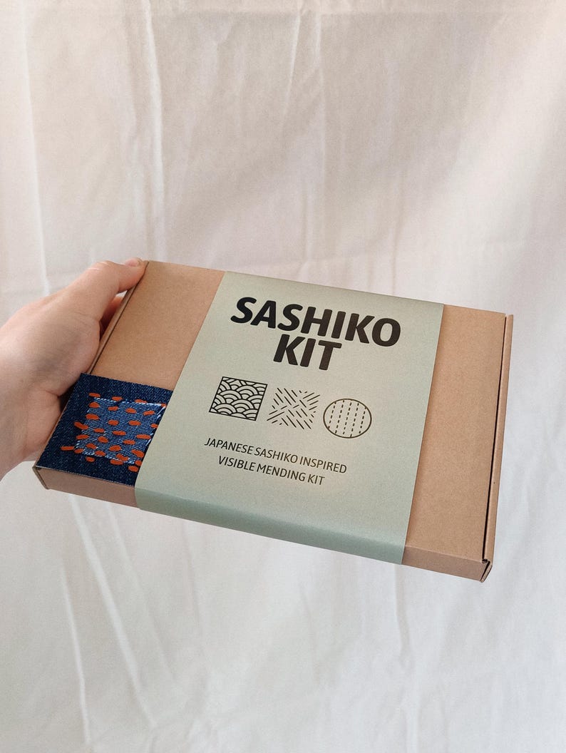 May include: A brown cardboard box with a green and white label that says "Sashiko Kit" and "Japanese Sashiko Inspired Visible Mending Kit". The label also has three small illustrations of different stitching patterns. The box is open and a blue fabric with orange stitching is visible inside.